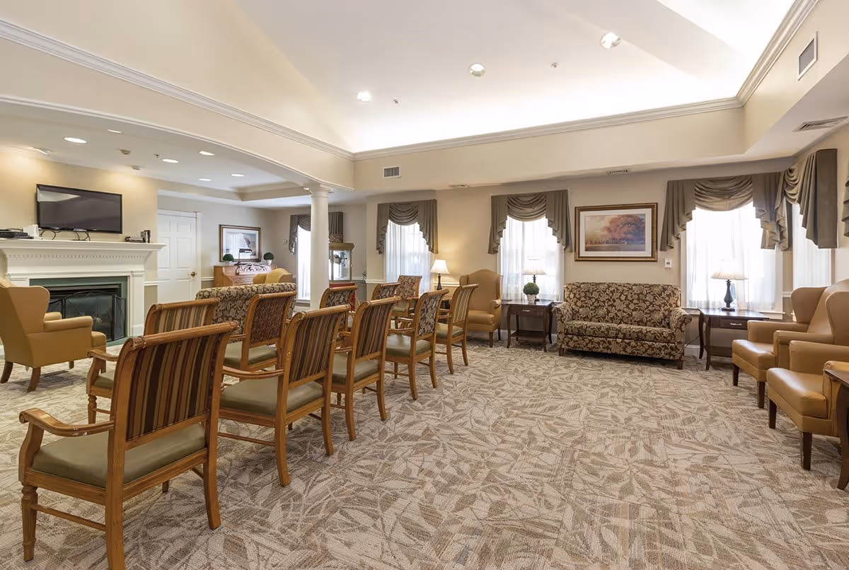 A spacious and well-lit living room area in Park Creek Place featuring multiple wooden chairs with cushions arranged in rows, patterned sofas, armchairs, side tables with lamps, a fireplace with a mounted TV above it, and large windows with draped curtains allowing natural light to fill the room.