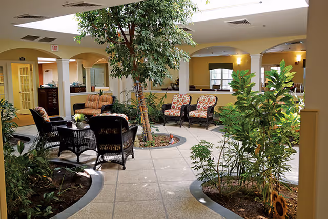 Indoor common area with a tiled floor and several seating arrangements including wicker chairs and a sofa with cushions. The space features a large indoor tree and various plants in built-in planters, with soft yellow walls and arched openings leading to other rooms.