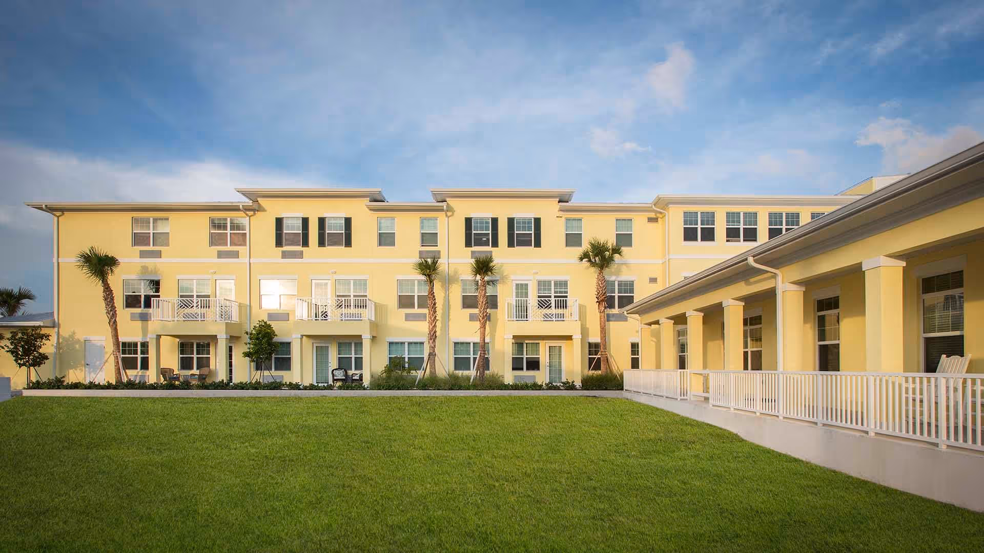Exterior view of a yellow three-story senior living community building with balconies, palm trees, and a well-maintained green lawn under a partly cloudy blue sky.