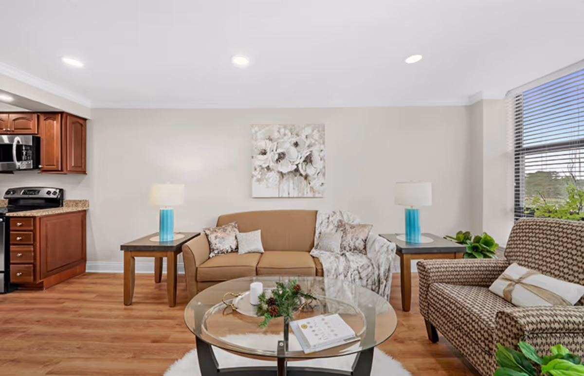 A cozy living room area with a brown sofa adorned with decorative pillows and a throw blanket, a patterned armchair with a pillow, two side tables each with a blue lamp, a glass coffee table with a candle and greenery centerpiece, and a kitchen area with wooden cabinets and stainless steel appliances visible on the left. The room has wooden flooring, a large window with blinds, and a floral painting on the wall.