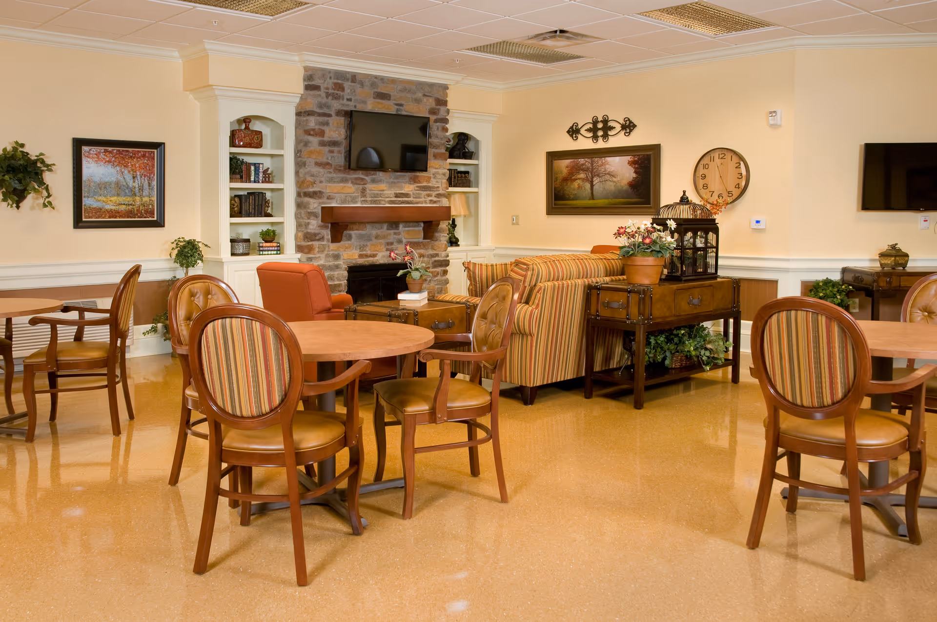 A cozy common area in a senior living facility featuring round wooden tables with striped cushioned chairs, a striped sofa, armchairs, a stone fireplace with a mounted TV above it, built-in white shelves with books and decor, framed artwork on the walls, a large clock, and various plants and decorative items.