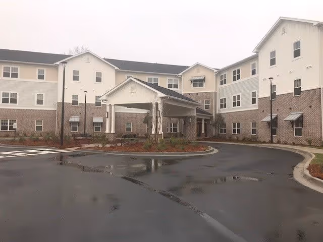Exterior view of a three-story senior living facility building with a covered entrance and a curved driveway in front. The building has a combination of brick and light-colored siding with multiple windows and some small awnings. The surrounding area includes some landscaping with small plants and mulch beds.