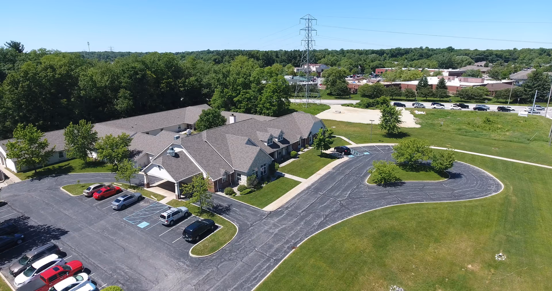 Aerial view of a senior living facility named Brookdale South Bend showing a single-story building with a gray roof surrounded by green lawns, trees, and a parking lot with several cars. The area is bordered by a road and other buildings in the distance under a clear blue sky.