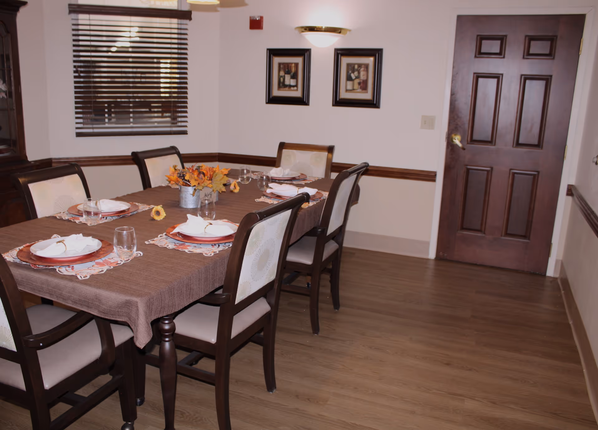 A dining room with a rectangular table covered with a brown tablecloth, set for six people with plates, glasses, and napkins. There is a small centerpiece with autumn-themed flowers. The room has wooden flooring, a window with blinds, two framed pictures on the wall, and a closed wooden door.