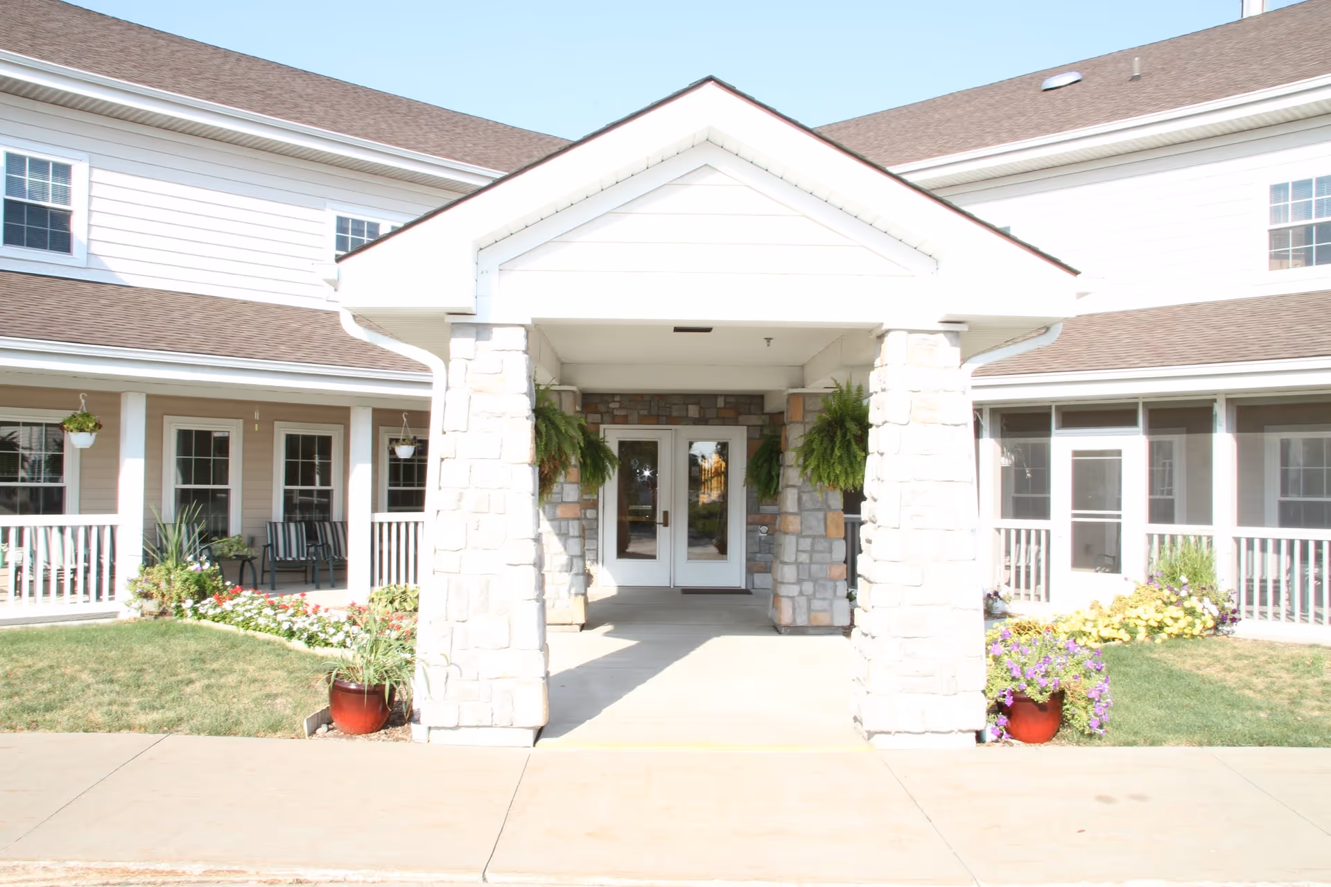 Front entrance of an assisted living building with a covered portico supported by stone pillars, double glass doors, hanging ferns, and potted flowers.