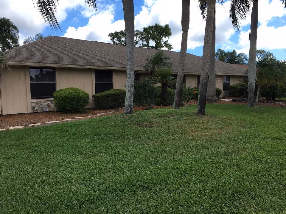 Single-story building with beige siding and a brown shingled roof, surrounded by green grass, palm trees, and shrubs under a partly cloudy blue sky.