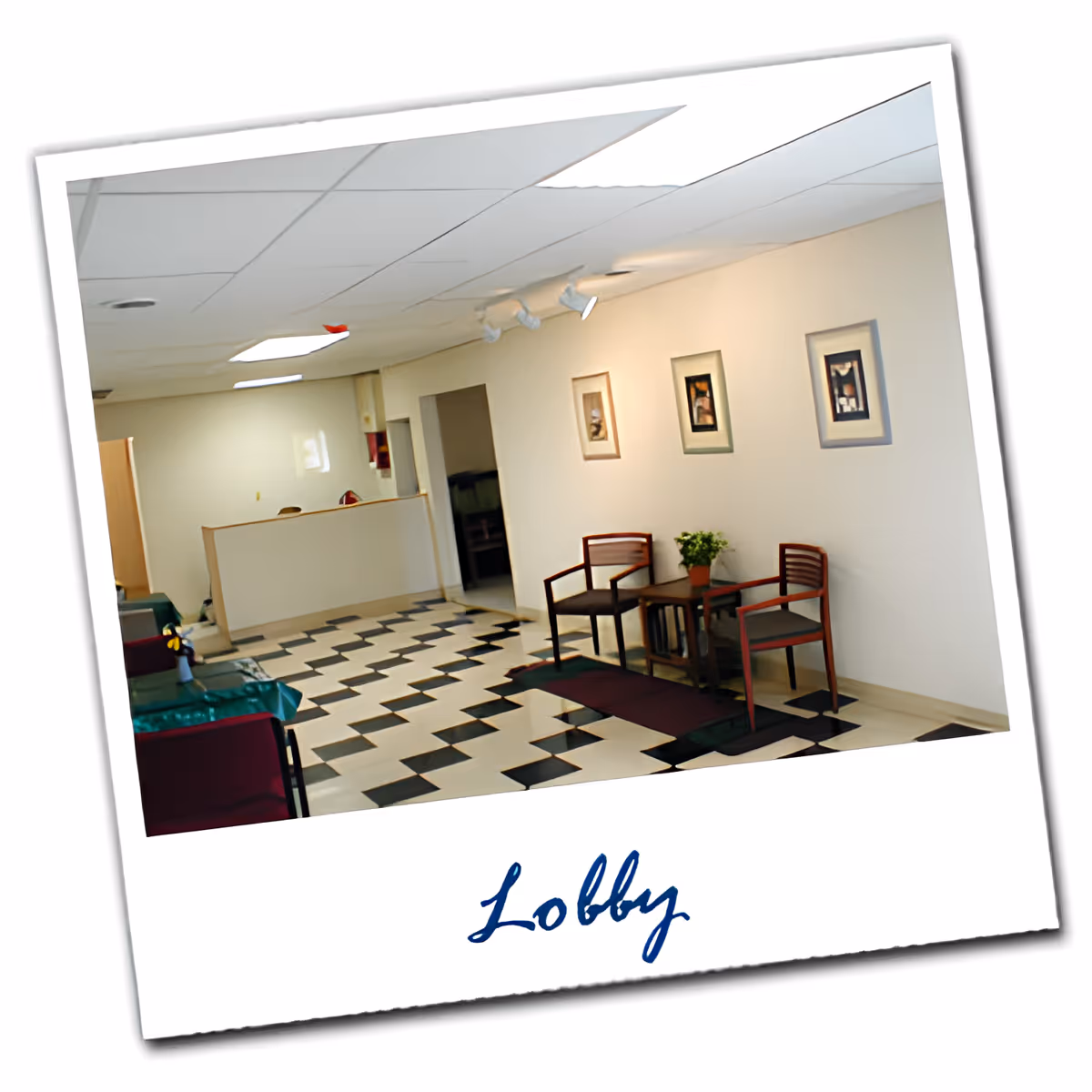 Interior view of a lobby area with a black and white checkered floor, two wooden chairs with cushions, a small table with a potted plant, three framed pictures on the wall, and a reception desk in the background.