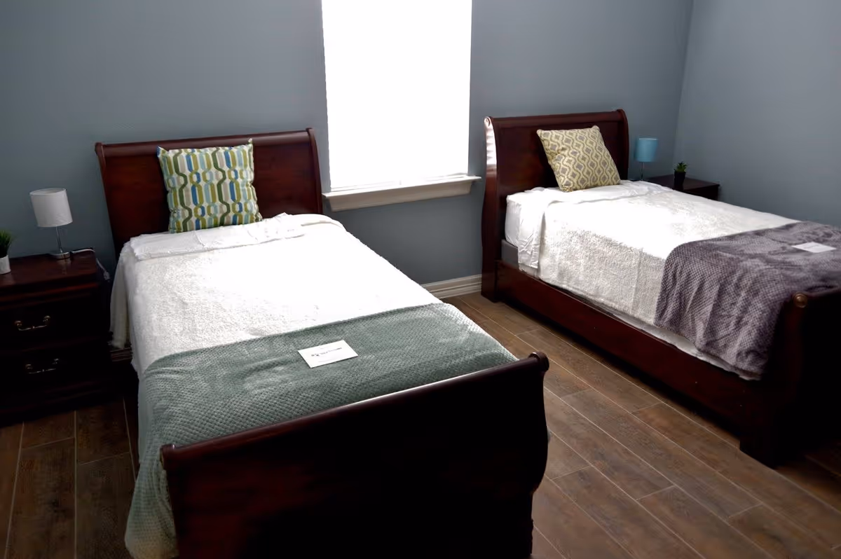 A bedroom with two single wooden beds, each with a white blanket and a decorative pillow. There is a window between the beds letting in natural light. Each bed has a nightstand with a small lamp and a plant. The floor is wooden, and the walls are painted light blue.