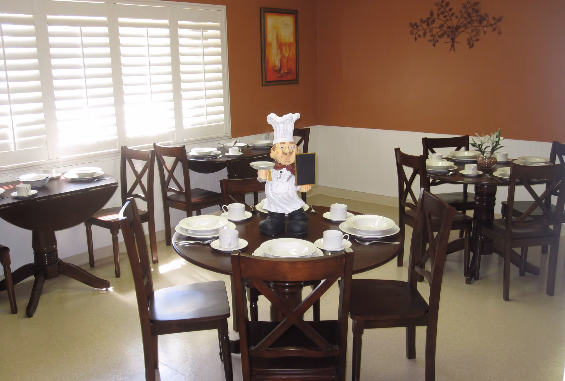 A dining room with several round wooden tables set with white plates, cups, and saucers. Each table is surrounded by wooden chairs with cross-back designs. One table has a chef figurine holding a small chalkboard in the center. The room has brown and white walls, a window with white shutters, and decorative wall art.