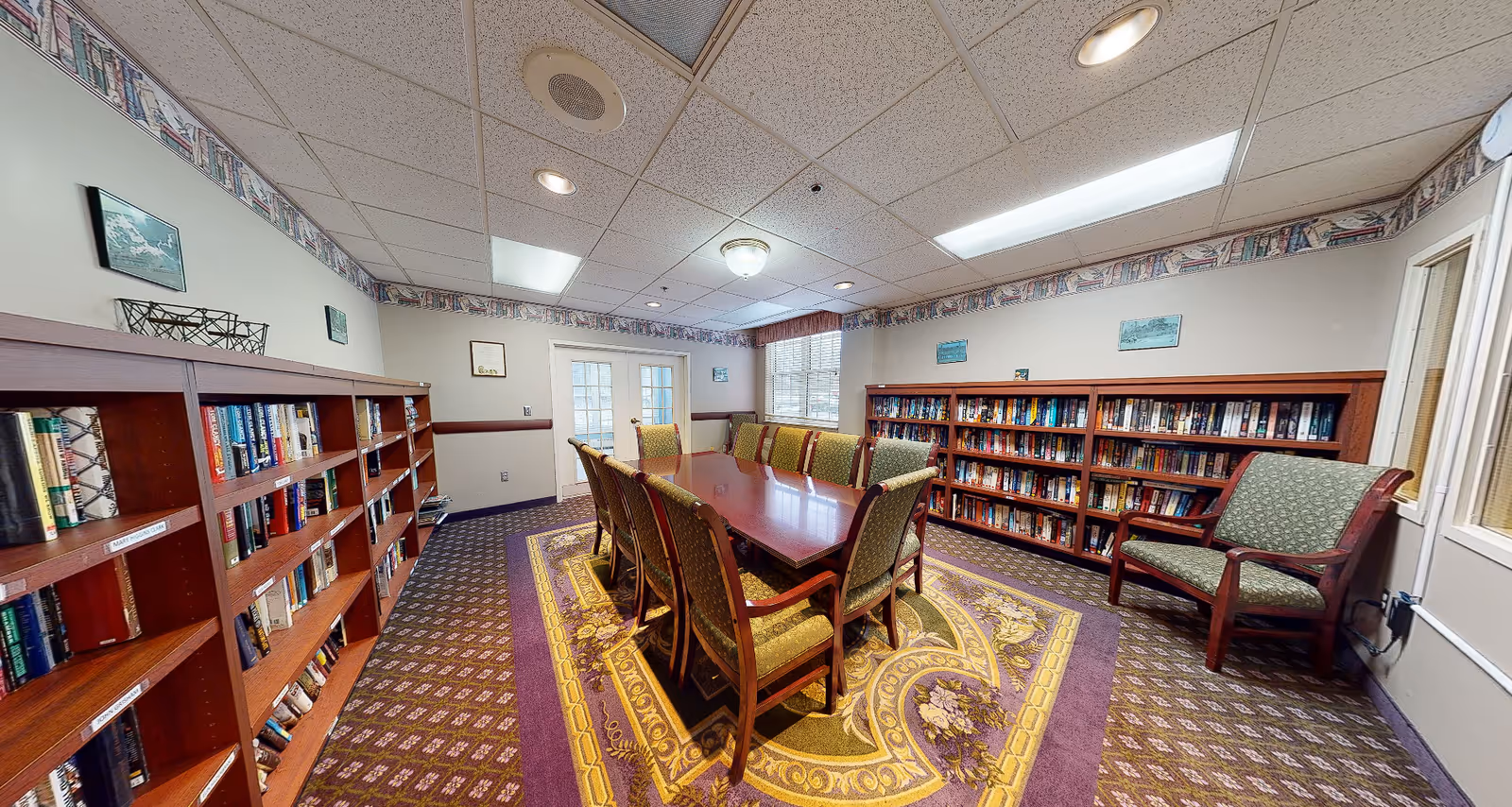 Interior community library/meeting room with a long table surrounded by chairs and bookshelves lining the walls.