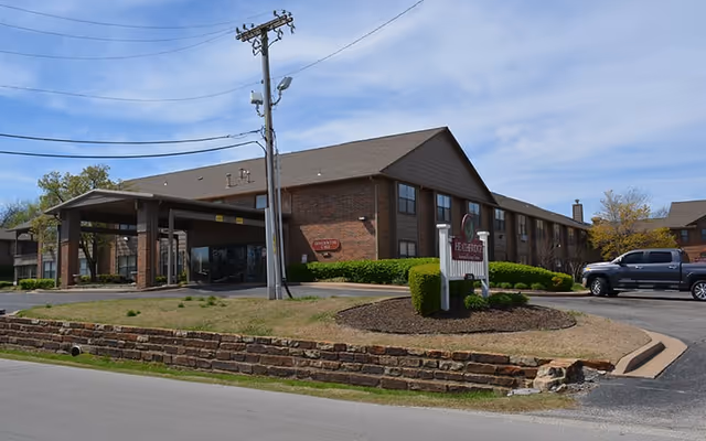Exterior view of Heatheridge Assisted Living & Residential Care Community building with a covered entrance, brick facade, and a sign displaying the facility's name. There is a pickup truck parked near the entrance and a utility pole with wires in front of the building.
