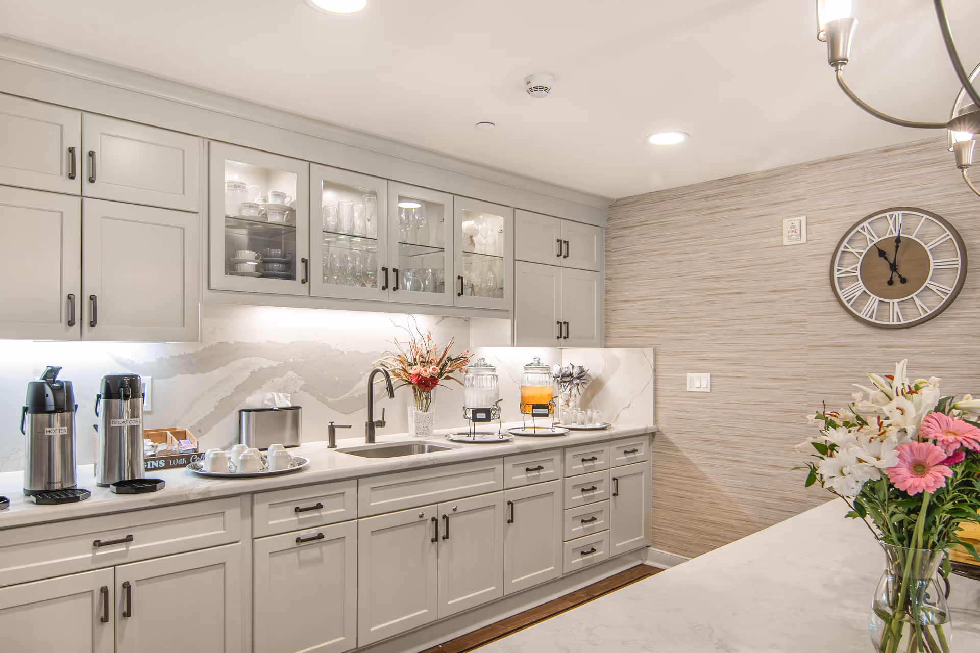 A bright and modern kitchen area with white cabinets and drawers, a marble countertop, and a marble backsplash. The upper cabinets have glass doors displaying cups and glasses. On the counter, there are two beverage dispensers, a vase with flowers, a tray with cups, and a coffee and tea station. A large wall clock is mounted on the textured beige wall, and a chandelier is partially visible on the right side above a countertop with a vase of flowers.