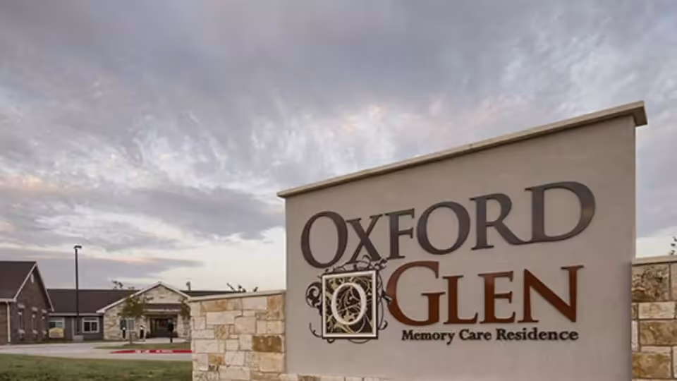 Exterior view of the Oxford Glen Memory Care Residence sign with the facility building and cloudy sky in the background.