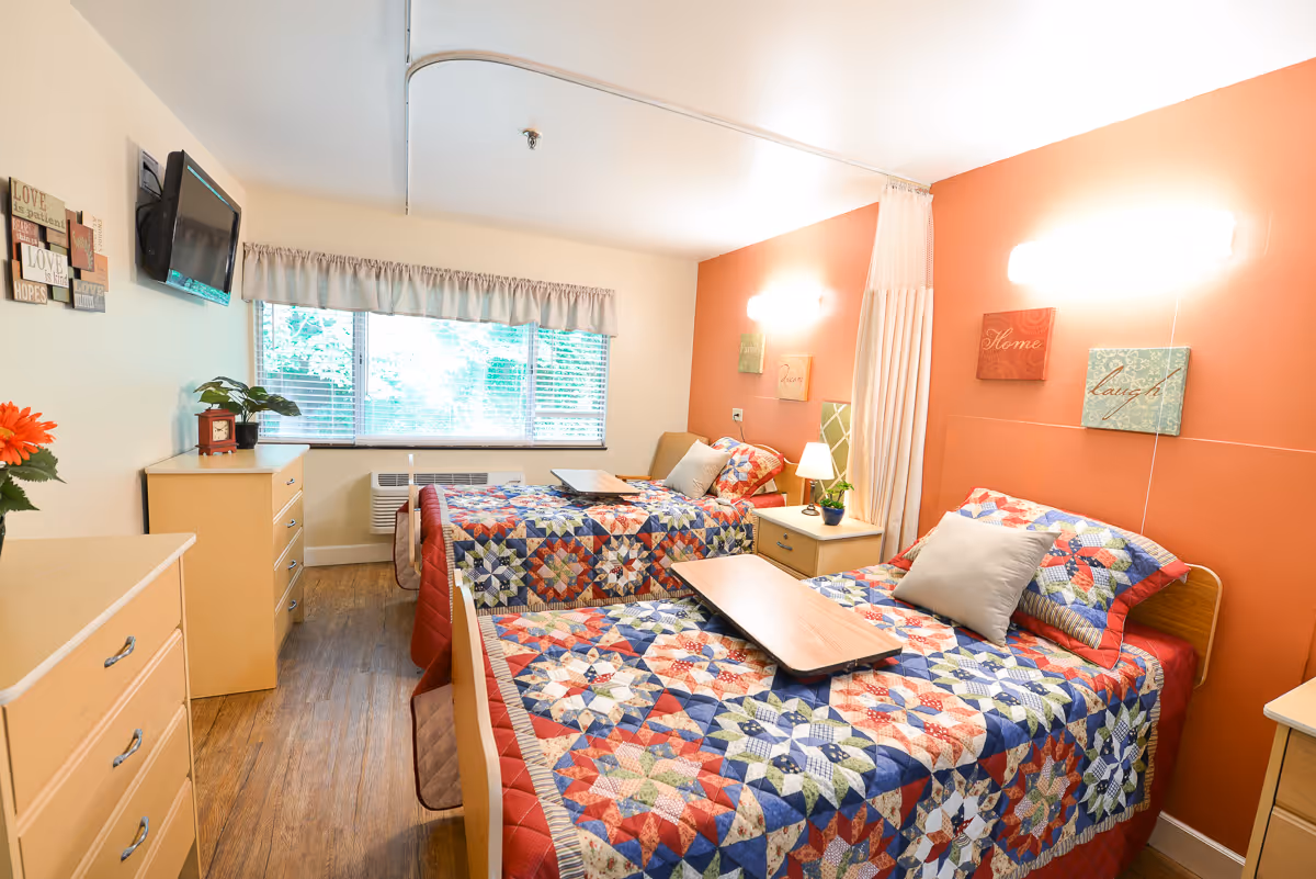 Two single beds with colorful patchwork quilts in a bright, neatly furnished senior facility bedroom with bedside tables, a TV, and a window.