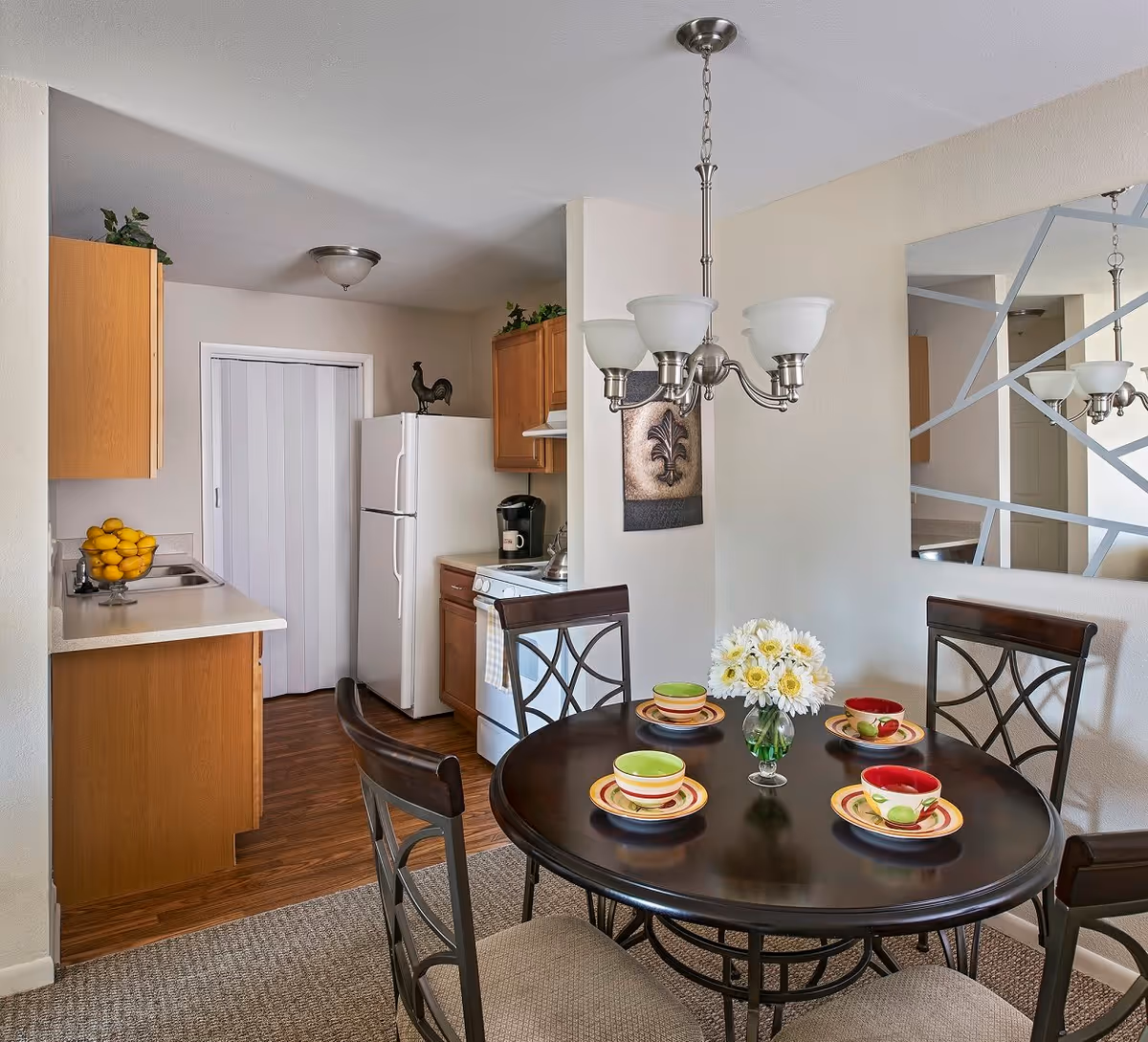 A cozy dining area with a round dark wooden table set with four colorful bowls and plates, a vase with white flowers in the center, and four chairs with cushioned seats. Adjacent to the dining area is a compact kitchen featuring wooden cabinets, a white refrigerator, a stove, a coffee maker, and a bowl of lemons on the counter. A modern chandelier hangs above the table, and a decorative wall mirror with geometric patterns reflects part of the room.