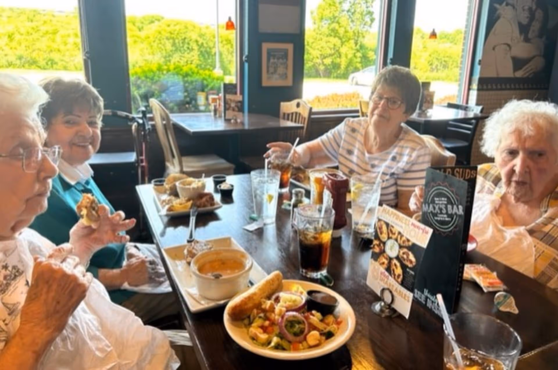 Four elderly women sitting around a table in a restaurant, enjoying a meal together with plates of food and glasses of drinks in front of them. The background shows large windows with a view of greenery outside.