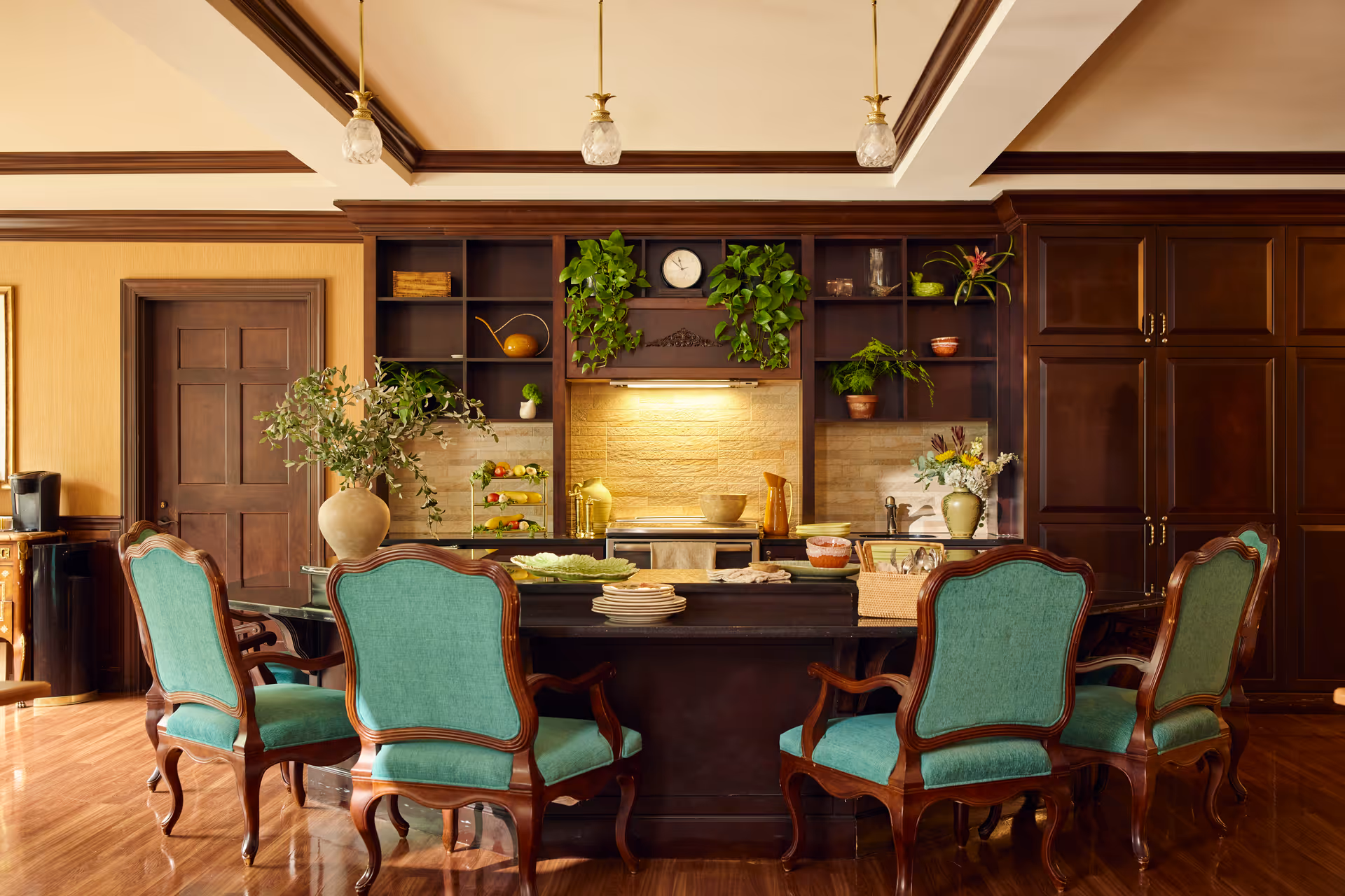 A warm, elegant kitchen and dining area featuring a dark wooden island with a black countertop surrounded by six vintage-style chairs upholstered in teal fabric. The background includes dark wood cabinetry, open shelves with decorative plants and pottery, a clock, and a stove with a lighted backsplash. The room has wooden flooring and a ceiling with recessed lighting and pendant lights.