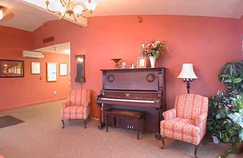 Pink-walled senior living lounge with piano, two upholstered chairs, plant, and lamp.