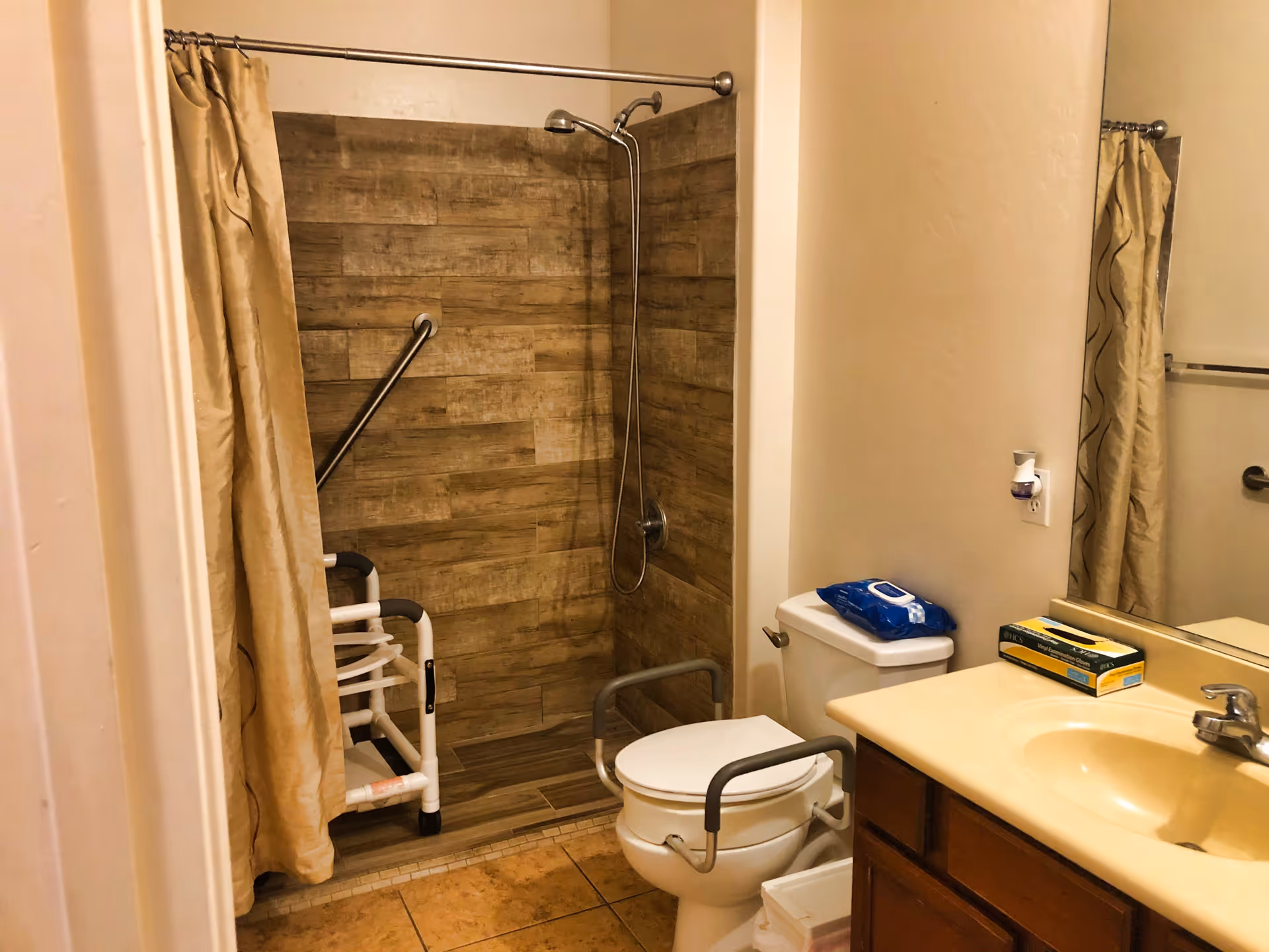 Accessible bathroom with a walk-in tiled shower, grab bars and shower chair, a toilet with support arms, and a sink with mirror.