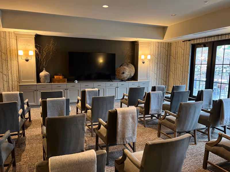 A cozy meeting or media room with multiple rows of gray upholstered chairs, each draped with a light gray throw blanket. The room features a large flat-screen TV mounted on a dark accent wall above a long cabinet with decorative vases and art pieces. Soft wall sconces provide warm lighting, and large windows with glass doors allow natural light to enter, showing an outdoor patio area.