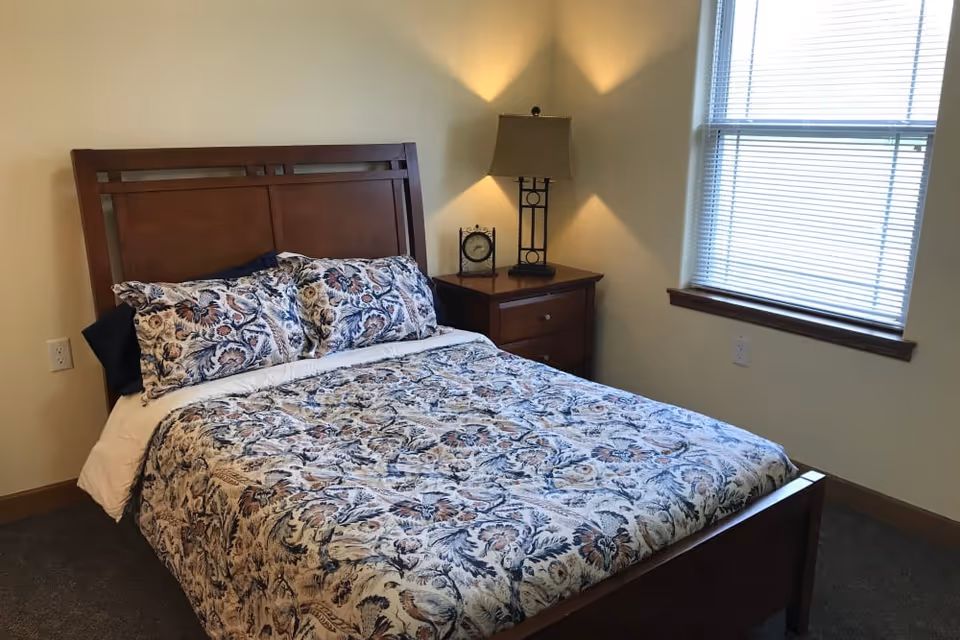 A bedroom with a wooden bed frame and a floral patterned bedspread and pillows. Next to the bed is a wooden nightstand with a decorative clock and a lamp. There is a window with blinds on the right wall, and the walls are painted a light beige color.