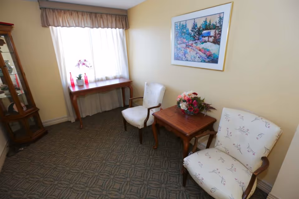 A small sitting area with two upholstered armchairs featuring floral patterns, a wooden side table with a flower arrangement, a wooden console table by a window with sheer curtains and valance, and a glass display cabinet in the corner. A colorful framed painting hangs on the wall above the side table.