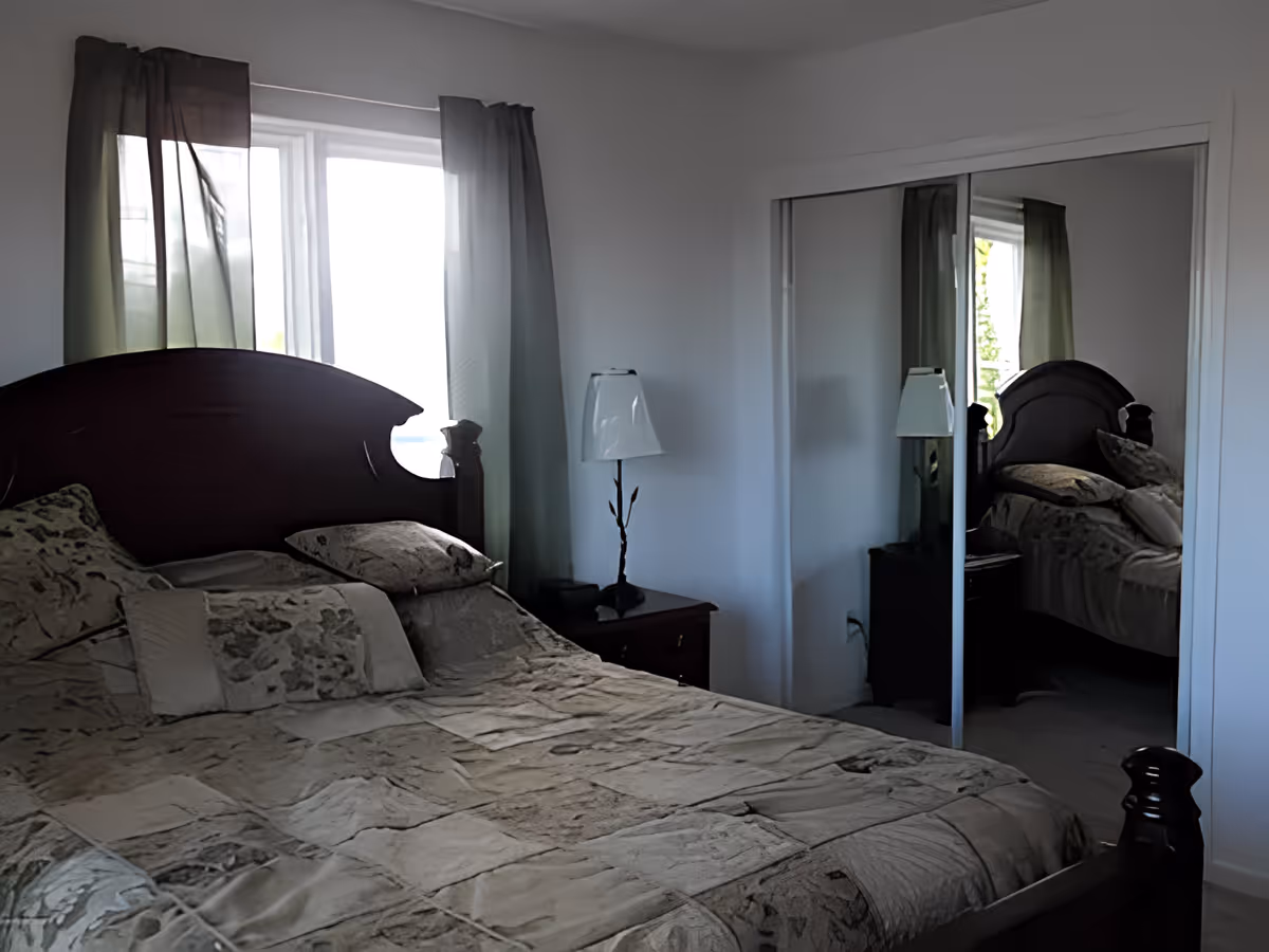 A bedroom with a large bed covered in a patterned beige and brown bedspread, two pillows, a wooden headboard, a nightstand with a lamp, and a window with sheer curtains. There is a mirrored sliding closet door reflecting the bed and window.