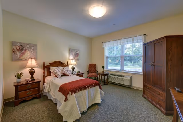 A cozy bedroom in a senior living facility featuring a single bed with white bedding and a red throw blanket, two wooden nightstands with lamps and decorative items, a wooden armoire, a window with white curtains, and a small seating area with a chair and side table.