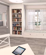 A bright interior room featuring a built-in bookshelf filled with books, a window with sheer white curtains, and a cushioned window seat with drawers underneath. In the foreground, there is a white table with a tablet placed on it. The floor has a wood-like finish.