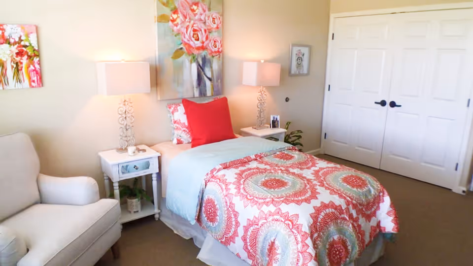 A bright and cozy bedroom featuring a single bed with a colorful red, white, and blue patterned bedspread and a red pillow. On each side of the bed are white nightstands with decorative lamps. There is a comfortable armchair to the left and floral artwork on the walls. A double-door closet is visible on the right side of the room.