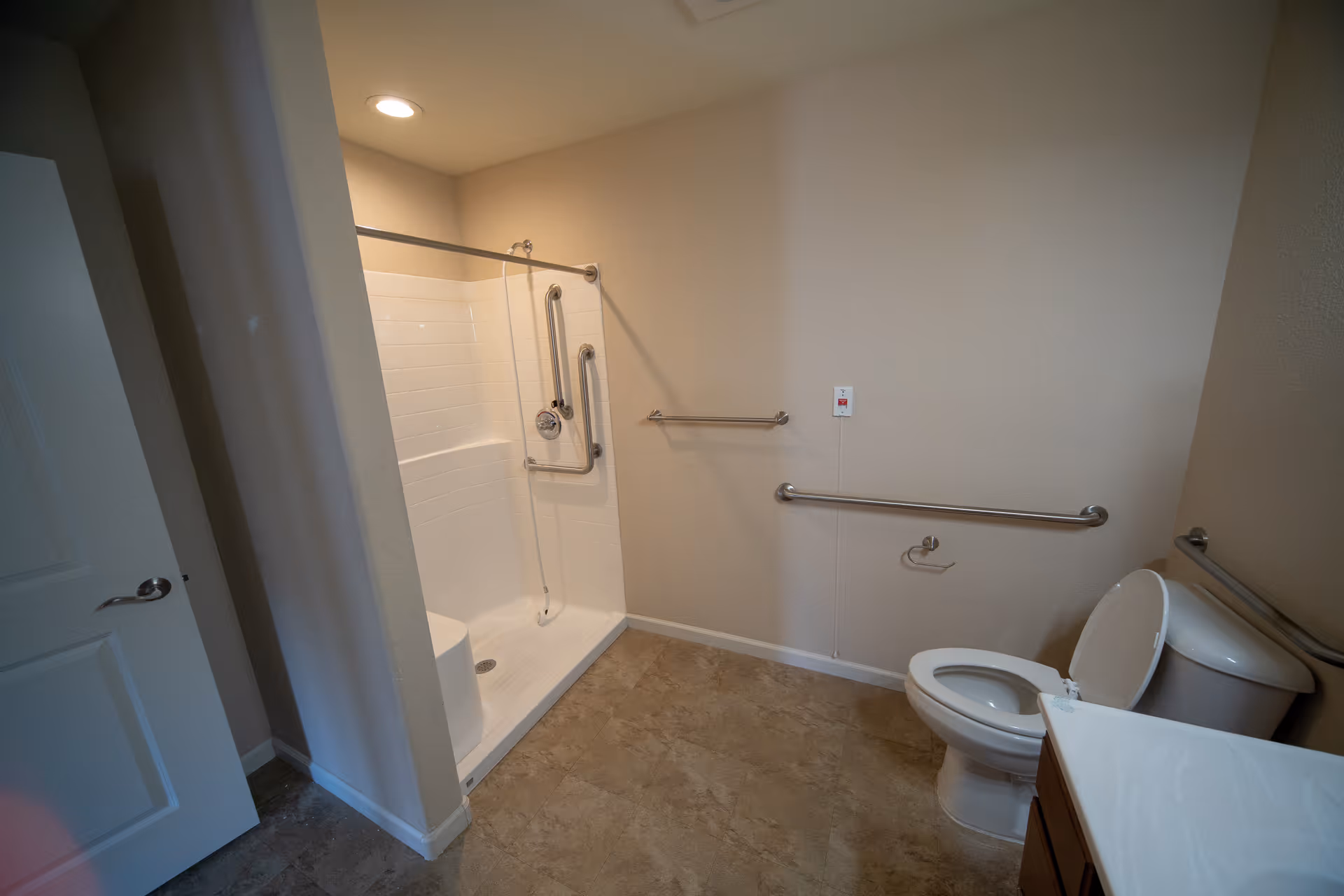 A spacious bathroom with a walk-in shower equipped with grab bars, a toilet with an open lid, and a vanity with a sink. The walls are beige, and the floor has a light brown tile. There are additional grab bars on the walls for accessibility and a pull cord emergency alert system.