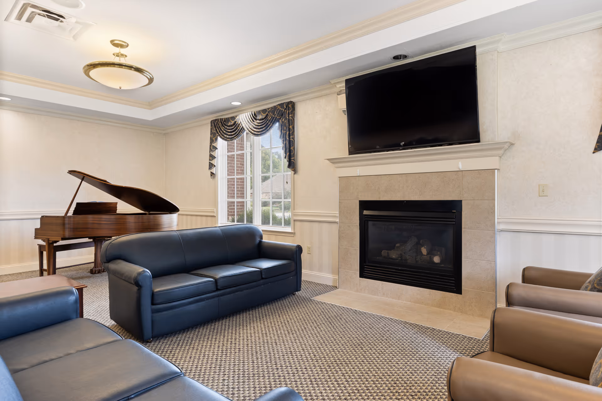 A cozy living room area with two dark blue leather sofas and two brown armchairs arranged around a beige tiled fireplace with a flat-screen TV mounted above it. A grand piano is positioned near a window with decorative curtains, and the room features a patterned carpet and soft lighting from a ceiling fixture.