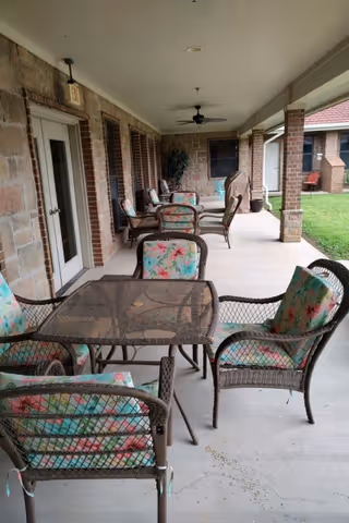 Covered outdoor patio area with multiple wicker chairs featuring floral cushions and glass-top tables. The patio is adjacent to a brick and stone building with ceiling fans and a view of a grassy lawn.
