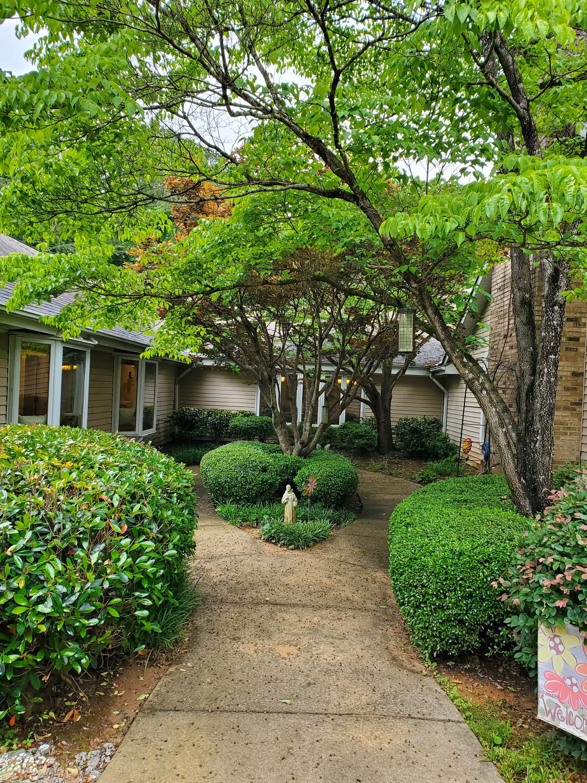 A paved walkway leading through a garden area with neatly trimmed bushes and trees, surrounded by building walls with windows. A small statue and decorative pinwheels are placed in the center of the garden bed. A colorful welcome sign is partially visible on the right side near the bushes.