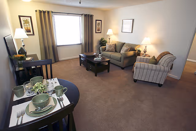 A cozy living room with a beige carpet, a green sofa with cushions, a striped armchair, a dark wooden coffee table, and two side tables with lamps. There is a window with curtains on the far wall and framed artwork on the walls. In the foreground, a small dining table is set with green dishes and silverware.