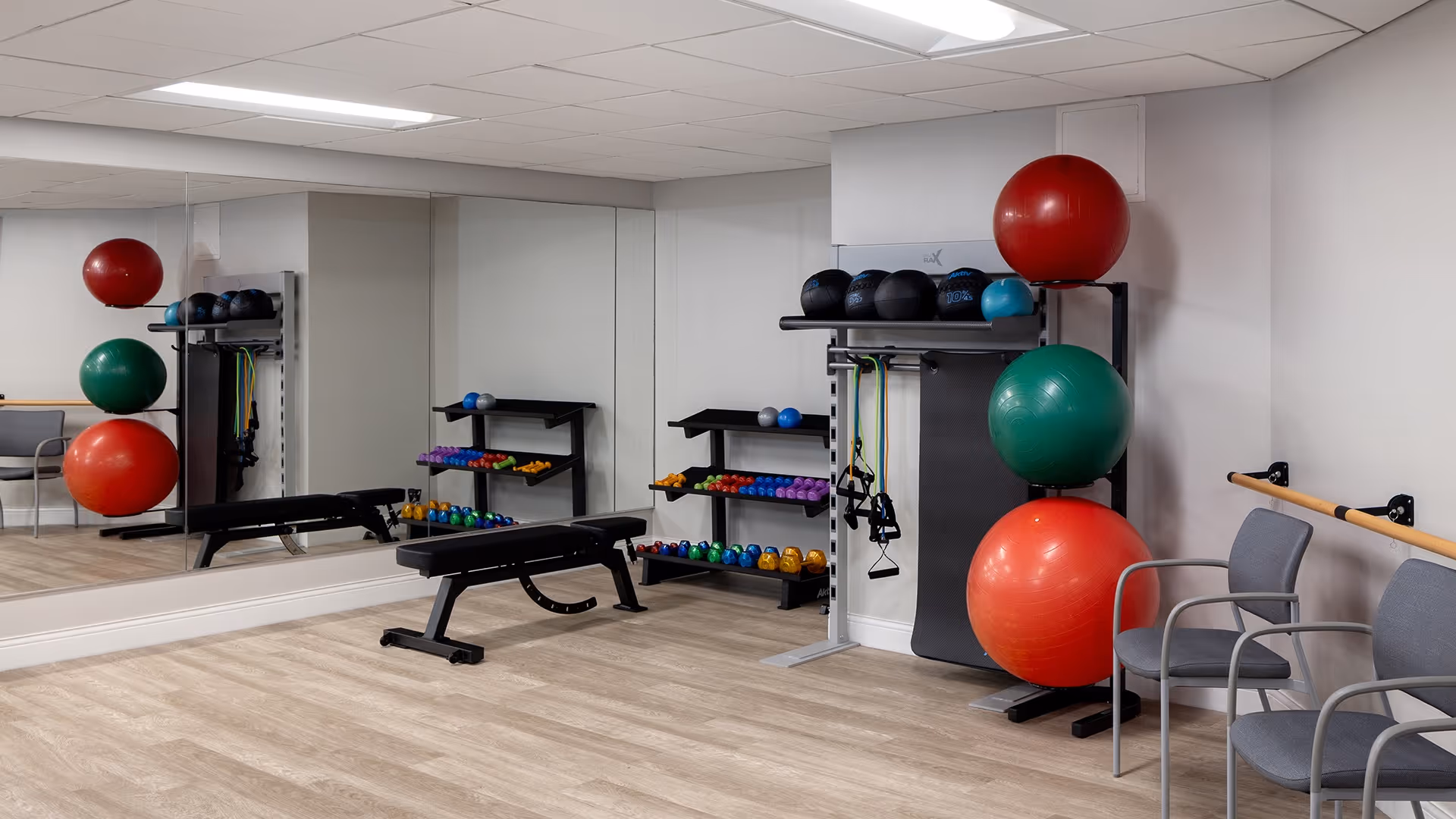 A small fitness room with a large wall mirror, a black workout bench, a rack holding colorful dumbbells, resistance bands, and three large exercise balls stacked vertically. Two gray chairs are placed against the wall with a wooden handrail behind them. The room has light wood flooring and white walls.