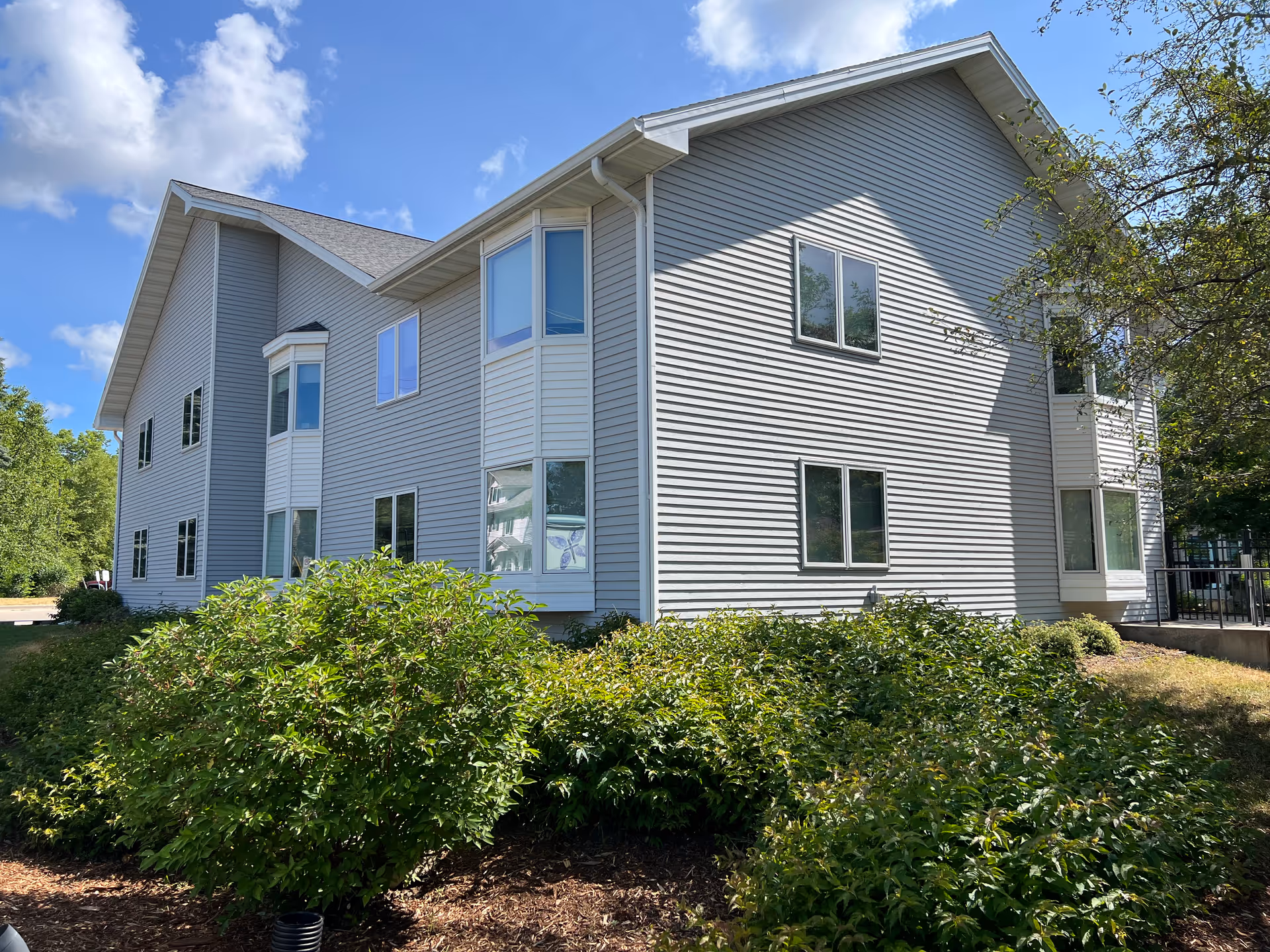 Exterior view of a two-story residential building with gray siding, multiple windows, and a sloped roof under a partly cloudy blue sky. Green bushes and trees surround the building.