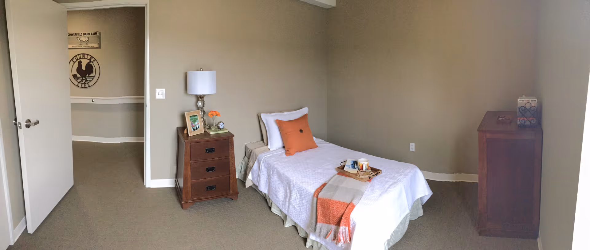 A simple bedroom with a single bed covered in white bedding and an orange throw blanket. There is an orange pillow on the bed and a wooden nightstand with a lamp, a framed photo, and a small flower vase. The room has beige walls and carpeted floor. A wooden dresser is placed against the right wall with a decorative item on top. The door to the room is open, showing a hallway with wall decorations.
