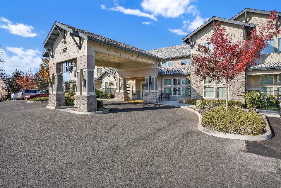 Exterior view of Almond Heights senior living facility entrance with a covered drop-off area, stone and beige siding facade, landscaped bushes and trees with red leaves, and a clear blue sky.