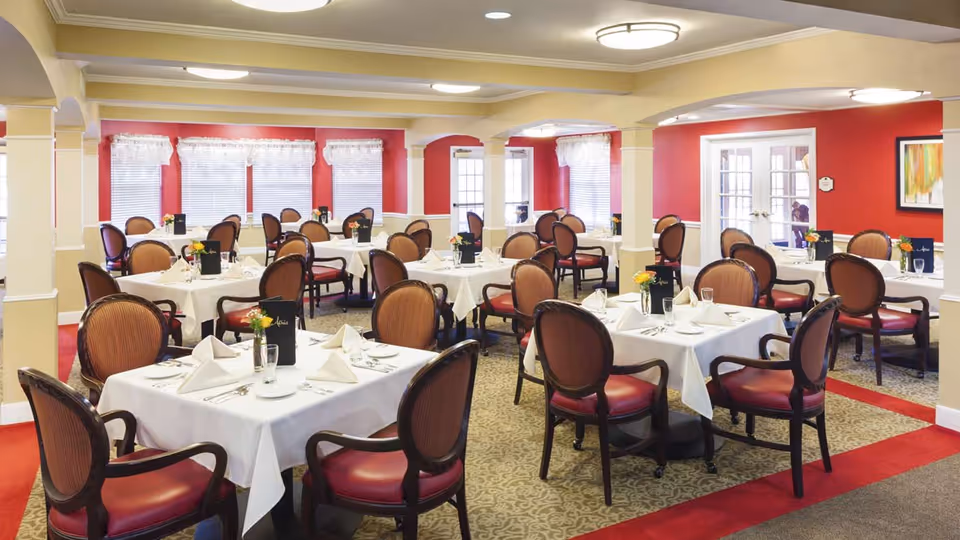 A well-lit dining room with multiple tables covered in white tablecloths, each set with folded napkins, glassware, and silverware. The chairs have wooden frames with red cushioned seats and backs. The walls are painted red with white trim, and there are windows with white curtains allowing natural light to enter. The room has a warm and inviting atmosphere.