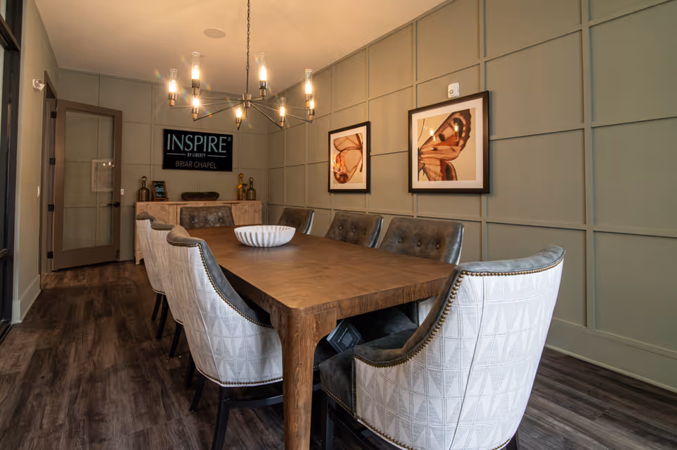 Long wooden dining table with upholstered chairs under a chandelier in a styled dining room with an "INSPIRE Briar Chapel" sign on the back wall.