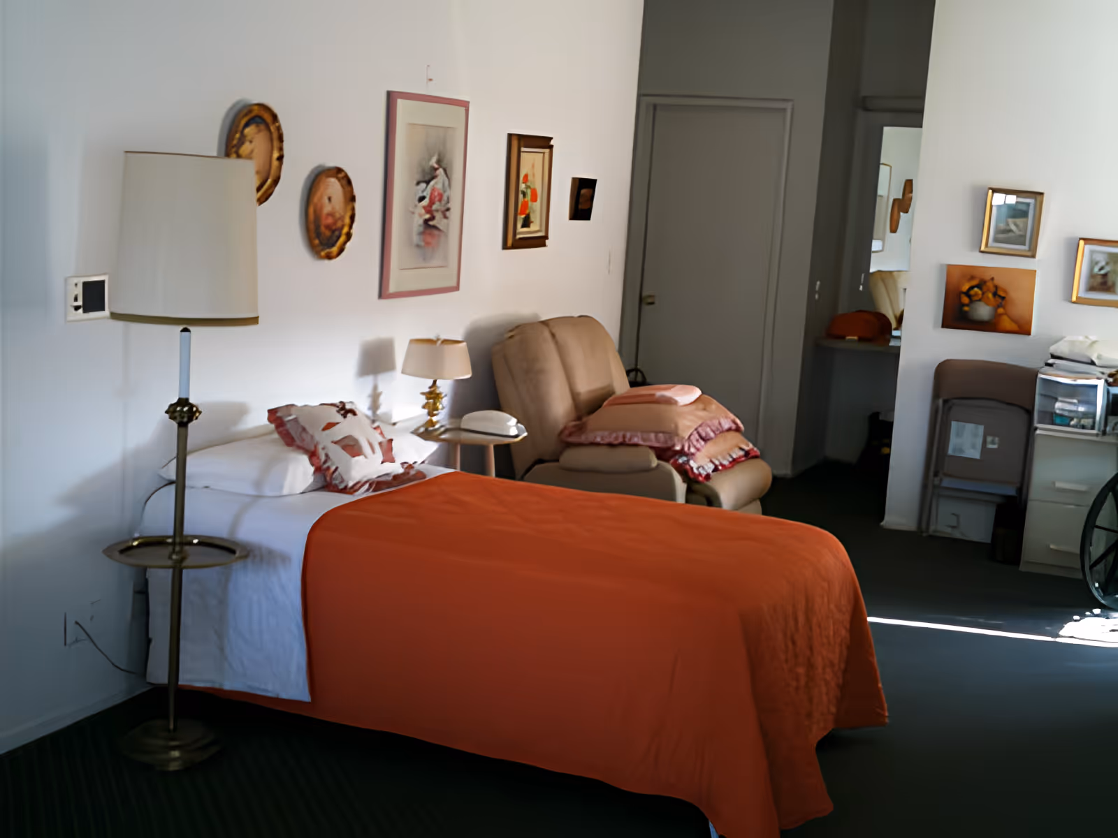 A bedroom with a single bed covered by an orange blanket, a floor lamp, bedside table, and a recliner with pillows against a wall decorated with framed artwork.