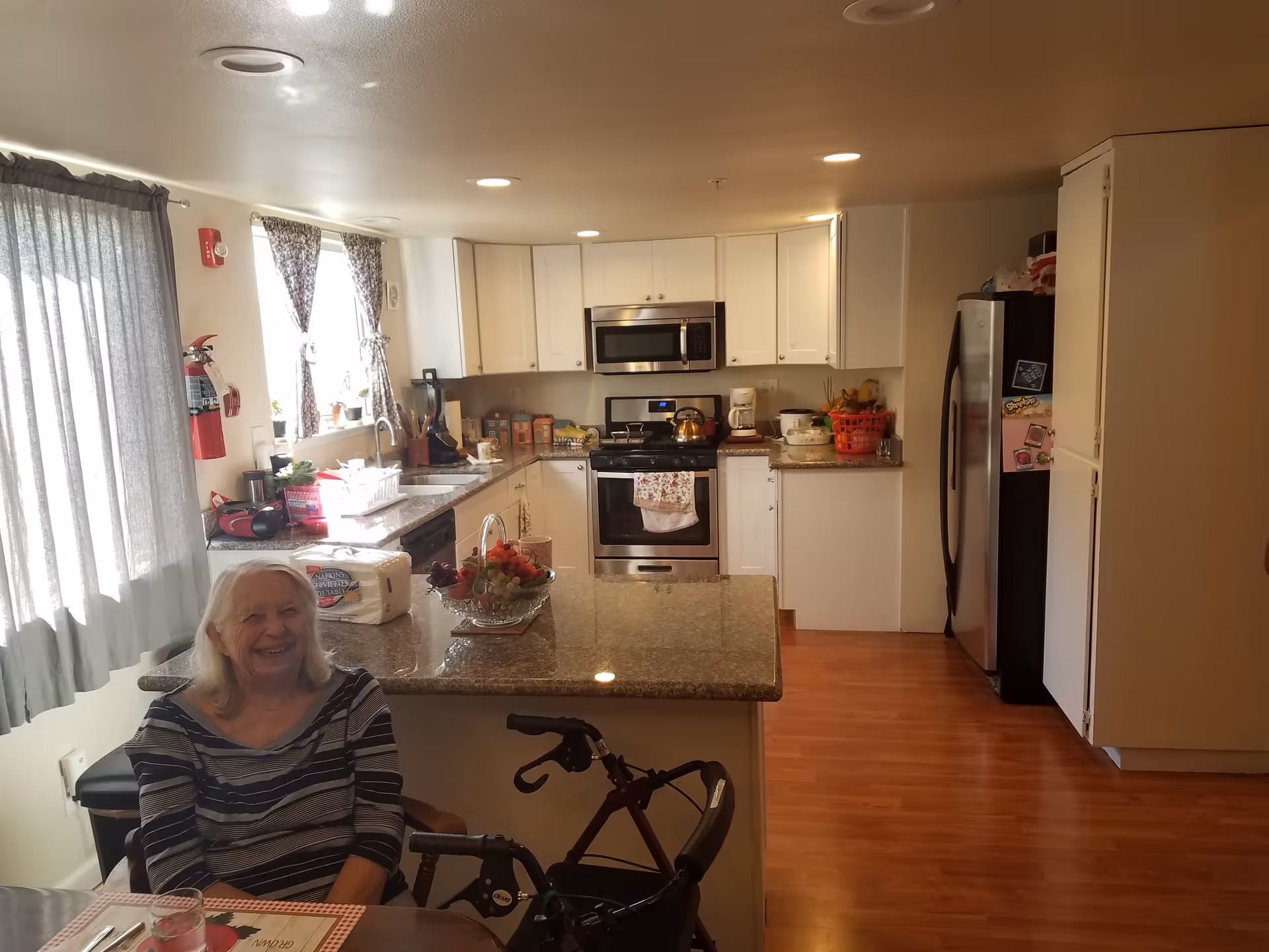 An elderly woman sitting at a table in a kitchen with wooden floors, white cabinets, and stainless steel appliances. The kitchen has a granite countertop island with a fruit bowl and paper towels on it. There are windows with curtains letting in natural light, and a walker is positioned near the woman.