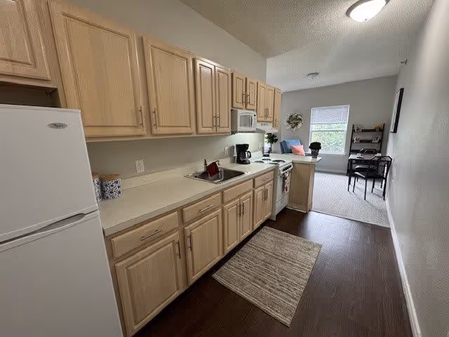 A compact kitchen area with light wood cabinets, a white refrigerator, microwave, coffee maker, and stove. The kitchen opens into a small living space with a window, a desk with a chair, and some decorative items including a wreath on the wall and a potted plant.