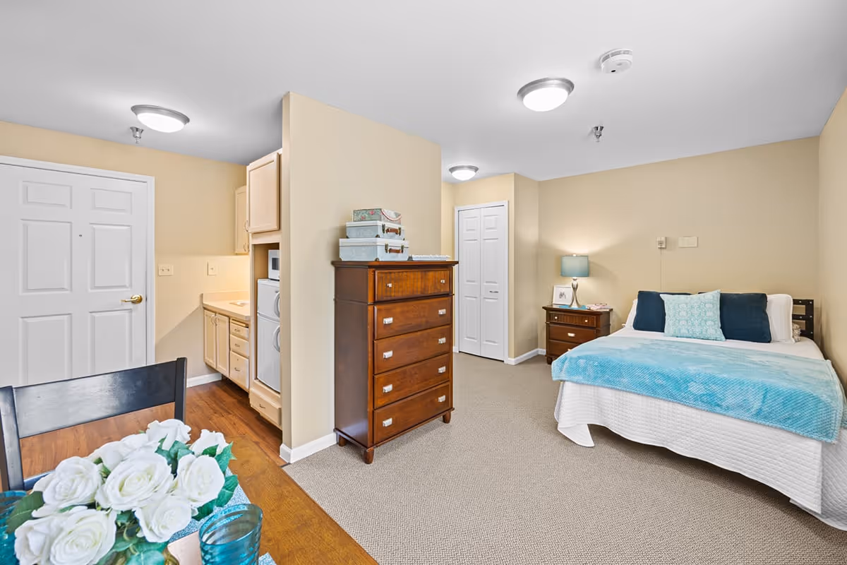 Bright studio apartment with a bed, wooden dresser, bedside table, small kitchenette and dining table with flowers.