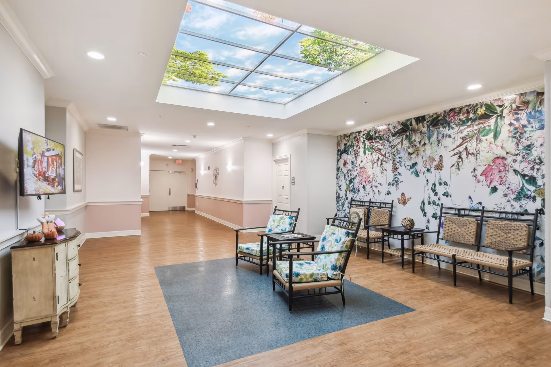 A bright and spacious hallway in a senior living facility with wooden flooring and a large skylight ceiling panel showing a blue sky with clouds and tree branches. The hallway features a seating area with floral cushioned chairs and woven benches arranged around small tables. One wall is decorated with a large floral mural, and a TV is mounted on the opposite wall above a small cabinet.