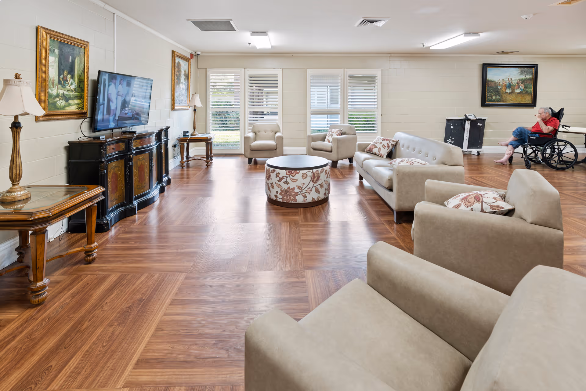 A spacious living room area in a senior living facility with beige armchairs and sofas arranged around a round floral ottoman. The room has wooden flooring, a TV mounted on a decorative cabinet, two table lamps, and framed paintings on the walls. A person in a wheelchair is sitting near the back wall, and large windows with white blinds allow natural light to enter.