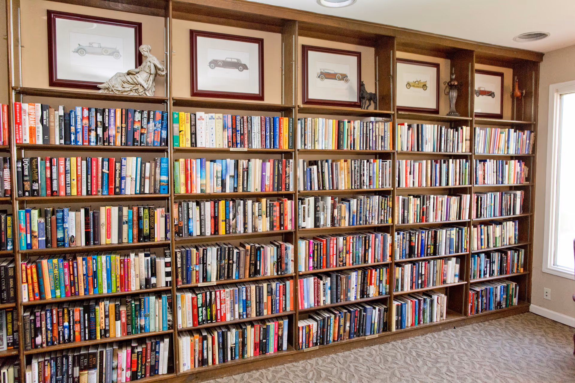 A large wooden bookshelf filled with numerous books arranged neatly on multiple shelves. Above the bookshelf are framed pictures of vintage cars and decorative sculptures. The room has a carpeted floor and a window on the right side allowing natural light in.