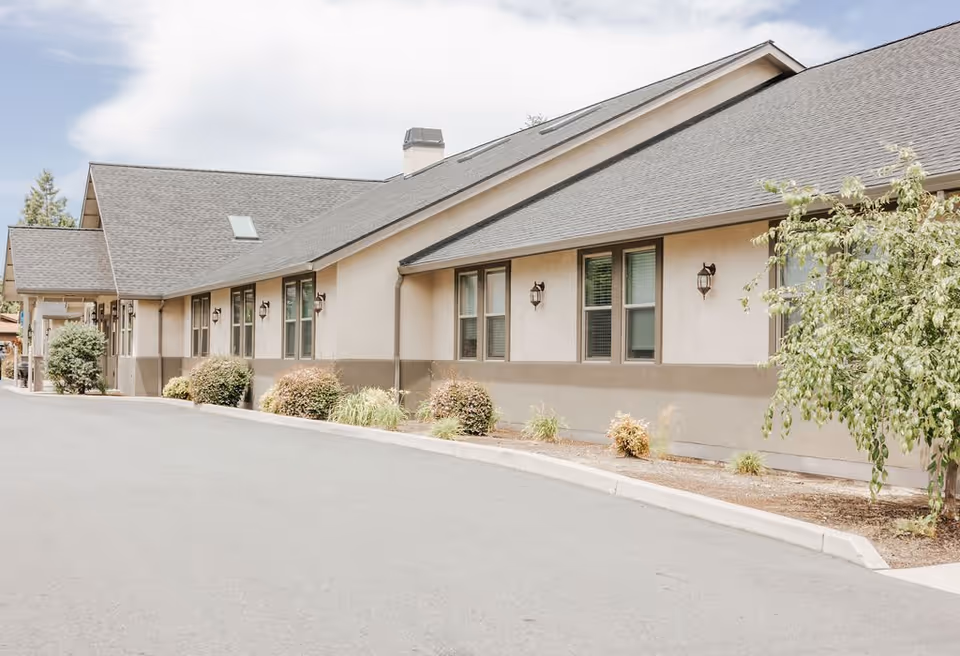 Exterior view of a single-story building with a sloped roof, multiple windows, and outdoor wall lights. There are small bushes and plants along the building's foundation and a paved driveway in front.
