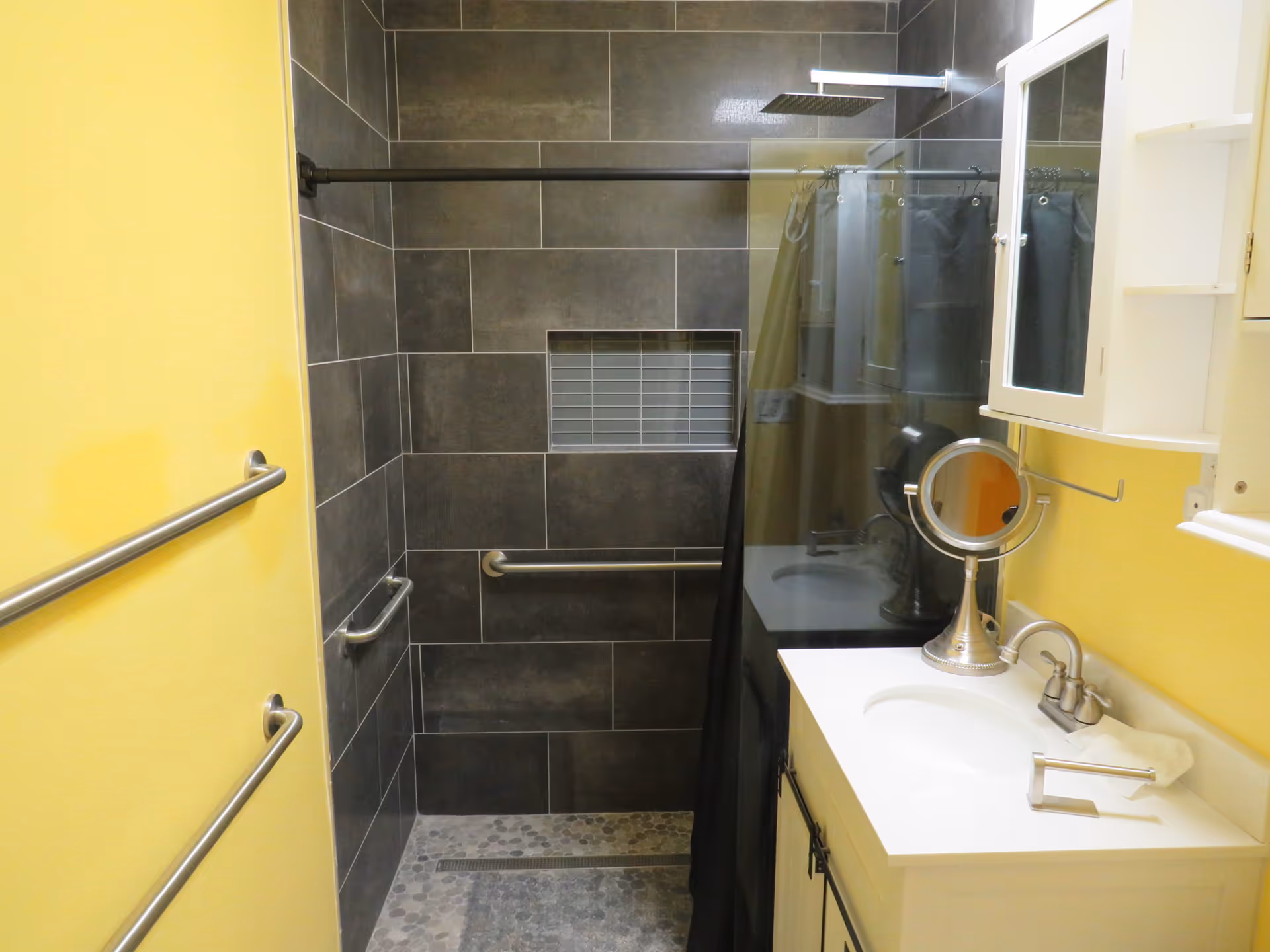 A bathroom with yellow walls and a walk-in shower featuring dark gray tiles and a built-in niche. The shower has a rainfall showerhead and grab bars. There is a white vanity with a sink, a faucet, a small round mirror, and a white cabinet with a mirrored door above it.